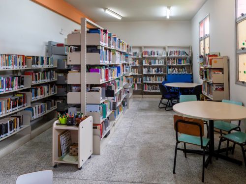 Sala de biblioteca com estantes repletas de livros e mesas redondas para leitura. Nas laterais, cadeiras escolares e janelas de vidro com adesivos circulares coloridos. A iluminação é branca e uniforme.
