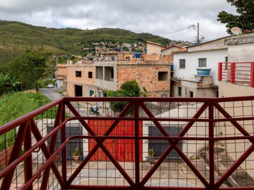 Vista da sacada com grade vermelha metálica para a rua. Em frente, casas e prédios inacabados de tijolos aparentes, além de vegetação e colinas verdes ao fundo. O céu está nublado.