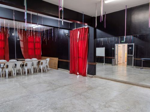 Sala ampla com piso de cimento, paredes pretas e espelho grande ocupando uma lateral. Do teto pendem fitas coloridas, e há cortinas vermelhas cobrindo janelas. Cadeiras brancas de plástico estão dispostas em fileira.