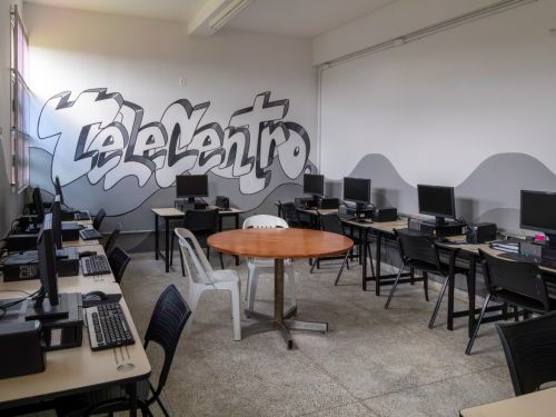 Sala de informática com fileiras de mesas equipadas com computadores de mesa, monitores e teclados. As cadeiras são de plástico preto e há uma mesa redonda de madeira no centro com cadeiras brancas e pretas ao redor. Na parede do fundo, há um grafite grande com a palavra “Telecentro” em letras estilizadas cinza e preto.