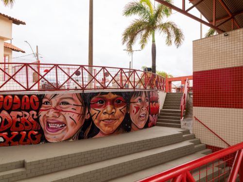 Escadaria externa com corrimãos vermelhos ao lado de parede com mural de três crianças indígenas pintadas com tintas faciais. Acima, guarda-corpo de grade vermelha e ao fundo, palmeiras e parte de um prédio com telhado cerâmico.