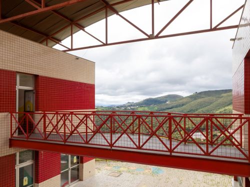 Passarela metálica suspensa de cor vermelha conecta dois blocos de prédio com fachada de pastilhas bege e vermelhas. Ao fundo, vista de colinas verdes sob céu nublado. Estrutura metálica do telhado visível acima da passarela.