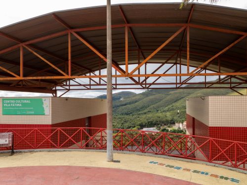 Área aberta coberta com telhado metálico e estrutura laranja. Na parede à esquerda, há uma placa verde com o texto “Centro Cultural Vila Fátima”. Embaixo, gradil vermelho e vista panorâmica de colinas cobertas por vegetação.