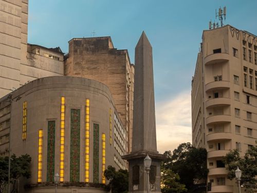 A Praça Sete de Belo Horizonte. No centro, destaca-se um obelisco alto de pedra, ladeado por luminárias de rua. À esquerda do obelisco, o Cine Theatro Brasil se ilumina, com sua fachada Art Déco e vitrais amarelos e laranjas brilhando. Ele está entre outros dois edifícios mais altos.