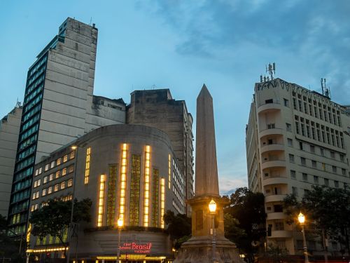 A Praça Sete de Belo Horizonte ao entardecer, com o céu já escurecendo e as luzes da cidade começando a brilhar. No centro, destaca-se um obelisco alto de pedra, ladeado por luminárias de rua. À esquerda do obelisco, o Cine Theatro Brasil se ilumina, com sua fachada Art Déco e vitrais amarelos e laranjas brilhando. Ele está entre outros dois edifícios mais altos.