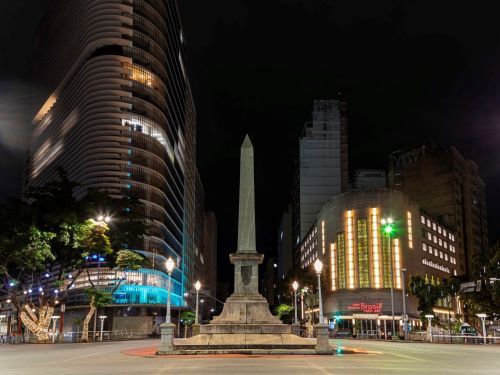 Foto noturna do obelisco da praça, visto de frente. À esquerda, um edifício moderno de fachada curva e branca, iluminado. À direita, o prédio histórico do Cine Theatro Brasil Vallourec, com suas janelas verticais iluminadas em tons de amarelo e verde. O céu está completamente escuro.