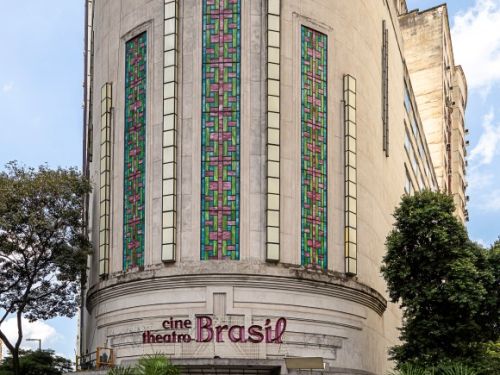 Edifício Cine Theatro Brasil, uma construção histórica em Belo Horizonte. No centro da foto, a fachada curva do prédio é visível. A entrada, ao nível da rua, possui grandes portas de vidro e metal. Acima da entrada, a fachada apresenta a inscrição "cine theatro Brasil" com letras de cor rosa, e detalhes arquitetônicos em relevo. Na parte superior da construção, grandes vitrais retangulares, com padrões coloridos em tons de verde, rosa e azul, se destacam. Em frente ao prédio, há palmeiras e pessoas.