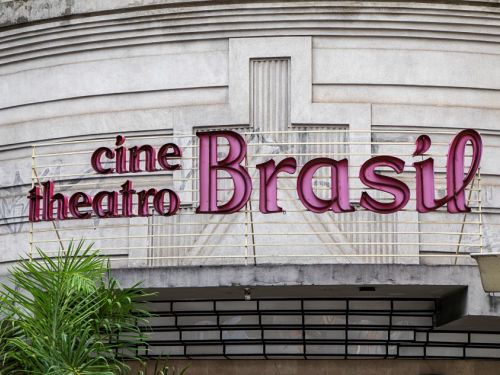 Um close da fachada do Cine Theatro Brasil, com foco na sua icônica sinalização. A placa tem letras de metal na cor rosa, com luzes internas que a fazem brilhar, e exibe as palavras "cine theatro Brasil". A parede do prédio, feita de concreto, apresenta linhas horizontais e detalhes em relevo que criam um desenho geométrico, característico da arquitetura Art Déco.