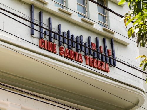 Um close da fachada do Cine Santa Tereza. A construção é de cor clara, com a parte inferior em um tom mais escuro. O nome "CINE SANTA TEREZA" está escrito em letras vermelhas de forma destacada. Acima do nome, há uma série de detalhes arquitetônicos verticais em azul marinho.