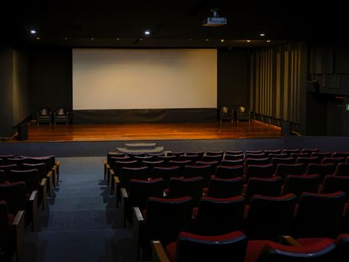 A parte da frente de uma sala de cinema ou teatro vazia. Em primeiro plano, as fileiras de poltronas vermelhas estão dispostas de forma ascendente, voltadas para o palco. No centro da cena, há um palco de madeira com degraus. Acima dele, uma tela de projeção grande e branca domina a parede escura.