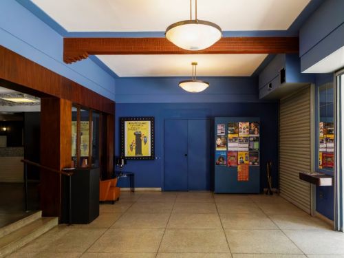 O hall de entrada do Cine Santa Tereza, que tem paredes pintadas em um tom de azul vibrante. O chão é coberto por azulejos claros e quadrados. No teto, pendem dois lustres de estilo vintage, com luminárias em forma de prato. Na parede do fundo, há um quadro grande com um pôster de filme e, ao lado, uma estante azul cheia de cartazes de eventos.