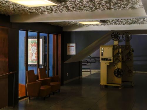 Uma sala de projeção e a área de espera do Cine Santa Tereza. Do lado direito, um projetor de filme antigo, de cor creme, com bobinas grandes, fica em destaque no chão. No teto, uma luz retangular ilumina o ambiente. Do lado esquerdo, é possível ver parte da área de espera. Há duas poltronas de couro marrom-claro, ao lado de uma parede com uma janela que revela um cartaz colorido. O teto tem um acabamento texturizado e áspero, parecendo bolhas ou uma esponja.