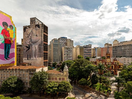 Vista urbana com prédios altos. Na lateral de um edifício, mural colorido mostra mulher negra de blusa vermelha, segurando uma criança no colo e com outra criança ao lado. Ao fundo, outro prédio com mural em tons claros retrata figura humana.