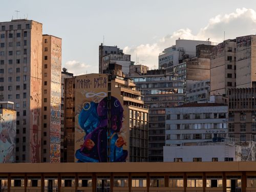 Paisagem urbana com prédios antigos e modernos. Em destaque, um edifício exibe mural colorido com duas figuras humanas de perfil, uma azul e outra roxa, rodeadas por peixes laranjas e símbolos cósmicos.