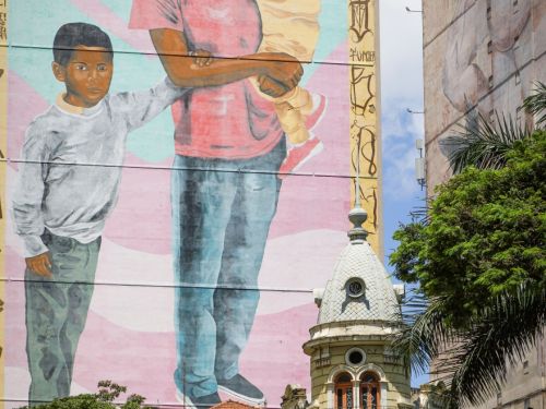 Mural em grande parede de prédio. Representa mulher negra de blusa rosa, segurando uma criança pequena no colo, e ao lado, um menino com camiseta clara. Fundo em tons claros com listras diagonais.