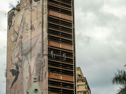 Prédio alto com fachada lateral ocupada por mural em tons marrons. A arte mostra figura humana de corpo inteiro, em posição inclinada, com pássaro preto ao lado. Parte da construção exibe janelas abertas sem vidros.