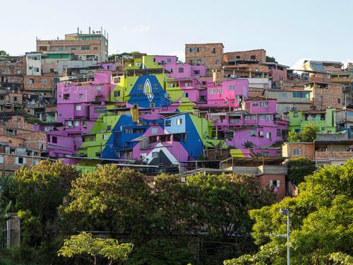 Conjunto de casas em morro. Muitas construções em tijolo aparente, algumas pintadas em cores vivas: rosa, azul e verde. As cores formam desenho geométrico que cobre várias fachadas.
