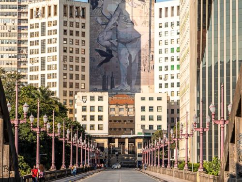 Ponte larga com postes de iluminação nas laterais. Ao fundo, mural em prédio alto retrata figura humana em tons claros, em posição inclinada, com pássaro preto. Cercado por prédios menores de várias janelas.