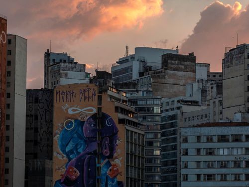 Cena urbana ao entardecer. Céu alaranjado atrás de prédios altos. Em destaque, fachada lateral com mural colorido mostrando duas figuras humanas de perfil, uma azul e outra roxa, com peixes laranjas em volta.
