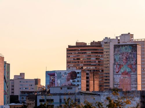 Conjunto de prédios no pôr do sol. Em destaque, mural em prédio cilíndrico mostra mulher de pele escura com saia laranja e bustiê verde, segurando folhas e espelho. Ao lado, outro mural com rosto de mulher negra de perfil, usando turbante vermelho.