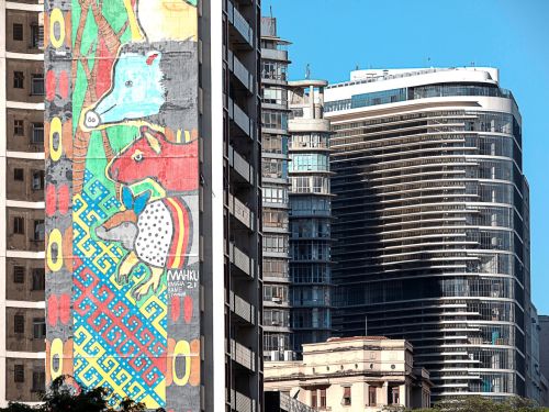 Mural colorido ocupa fachada lateral de prédio. A arte mostra animais estilizados, incluindo anta azul, boi vermelho e ave com corpo branco e pintas pretas, sobre fundo com padrões geométricos coloridos. À frente, prédio curvo de vidro moderno.