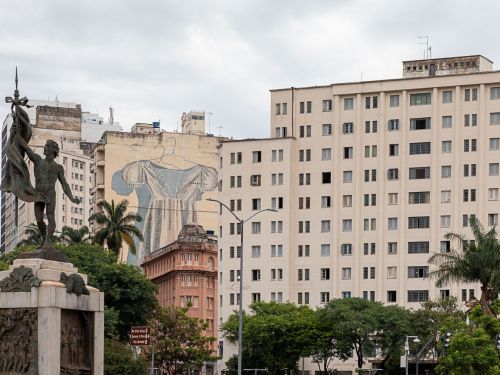 Vista urbana com escultura em primeiro plano e edifícios ao fundo. Um dos prédios tem um mural de arte representando uma figura com vestido azul. Céu nublado e presença de árvores na cena.
