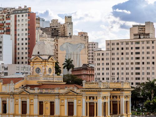 Imagem mostra um conjunto de prédios no centro da cidade com destaque para uma construção amarela com cúpula e relógios. Ao fundo, há um mural de arte com figura usando vestido azul claro em um prédio alto.