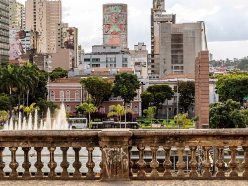 Vista de uma praça com chafariz, balaustrada de pedra e área pavimentada em primeiro plano. Ao fundo, diversos prédios com murais artísticos, incluindo um com retrato colorido de uma mulher segurando flores.