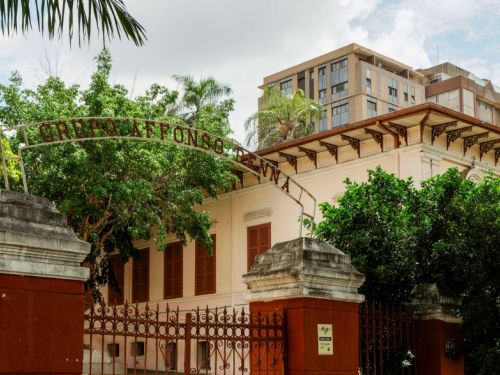 Fotografia da fachada de uma escola histórica cercada por portão de ferro trabalhado em vermelho-escuro. O muro baixo, na mesma cor, é encimado por pilares de pedra. Um arco metálico com o nome da instituição marca a entrada principal. O prédio, em estilo clássico, tem paredes claras, janelas de madeira em tom terroso e detalhes ornamentais na cornija do telhado. Árvores frondosas e palmeiras emolduram a construção, enquanto ao fundo se destacam edifícios modernos, criando contraste entre p
