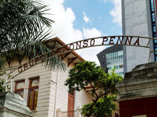 Entrada da Escola Afonso Pena a partir de um ângulo levemente inclinado para cima. Em primeiro plano, destaca-se o arco metálico com o nome da instituição em letras vermelhas. Logo atrás, parte da fachada do edifício histórico é visível, com paredes claras e janelas de madeira emolduradas por detalhes decorativos. Árvores e uma palmeira compõem o cenário, enquanto ao fundo surgem prédios altos e modernos, criando um contraste marcante entre o patrimônio histórico escolar e a vertic