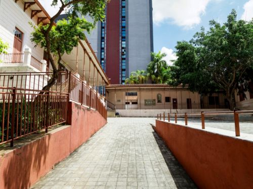 A fotografia mostra uma área externa pavimentada que conduz a um prédio escolar de arquitetura antiga, com fachada clara, janelas e portas de madeira. À esquerda, um muro baixo avermelhado com gradil acompanha a cena. No centro, o caminho leva a construções térreas com telhado colonial e detalhes em madeira. À direita, árvores altas oferecem sombra e compõem o ambiente. Ao fundo, ergue-se um edifício moderno que contrasta com o conjunto histórico.