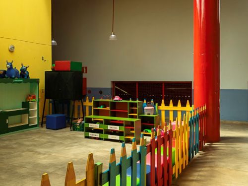 Área interna ampla com pé direito alto, paredes amarelas e um pilar cilíndrico vermelho. Em primeiro plano, uma cerca baixa de madeira, com estacas pintadas em várias cores vibrantes, delimita um espaço com brinquedos e estantes baixas e coloridas sobre um piso de cerâmica clara.