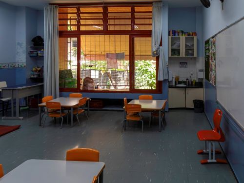 Sala de aula com paredes azuis e piso cinza. Há duas mesas infantis com cadeiras laranjas no centro. Ao fundo, uma grande janela com esquadrias vermelhas e grades amarelas. No canto direito, uma pequena pia com armário e um quadro branco na parede. A iluminação é suave.