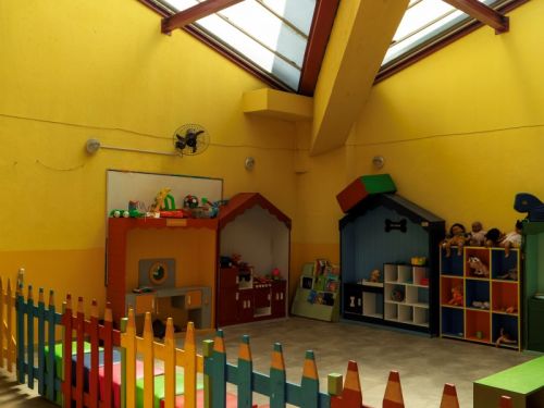 Espaço de recreação interno com paredes amarelas e teto com claraboia que permite a entrada de luz natural. Uma cerca baixa, com estacas em formato de lápis de cor, está em primeiro plano. Ao fundo, há casinhas de brinquedo, estantes com bichos de pelúcia e um pequeno teatro infantil.