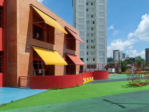 Área externa da escola com gramado sintético verde e piso azul. A fachada é de tijolos aparentes, com janelas protegidas por toldos de lona, sendo três amarelos e um vermelho. Ao fundo, um alto edifício residencial branco se destaca contra o céu azul com algumas nuvens.