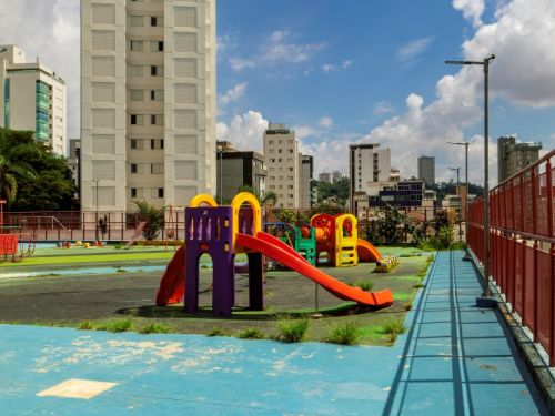 Parquinho externo em um terraço com piso azul. Um brinquedo de plástico colorido com escorregador vermelho está no centro. À direita, uma rampa de acesso com piso metálico e grade vermelha. Ao fundo, a paisagem urbana, com um grande prédio branco e outros edifícios sob um céu azul.