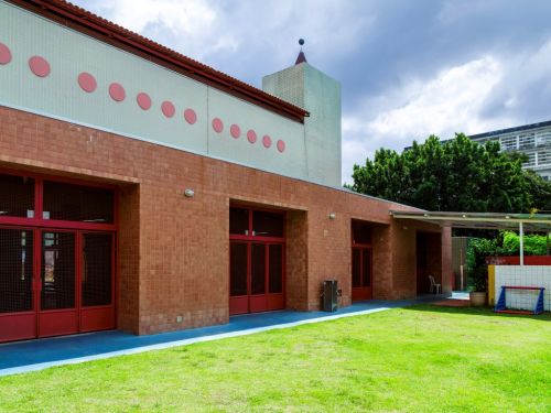 Fachada da escola vista a partir de um gramado verde bem aparado. A base do prédio é de tijolos laranjas, com portas e janelas de esquadrias vermelhas. A parte superior é branca com círculos decorativos cor-de-rosa. Ao fundo, árvores e um céu nublado.