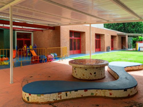 Pátio coberto com piso laranja. No centro, um banco de concreto de formato orgânico e uma mesa redonda, ambos revestidos com mosaicos. Ao fundo, um parquinho com brinquedos coloridos, cercado por uma grade amarela. A fachada de tijolos aparentes e um gramado verde completam a cena.