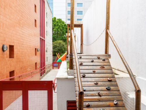 Um corredor estreito entre um muro branco à direita e um prédio de tijolos à esquerda. Em primeiro plano, uma rampa de escalada de madeira com agarras coloridas e rede de proteção. Ao fundo, um portão de metal vermelho e a continuação do corredor, com um prédio alto visível ao longe.