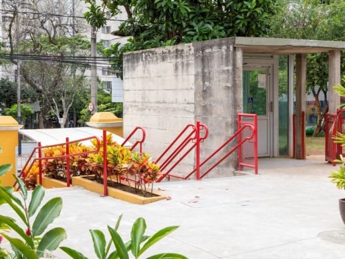 Entrada da escola com uma rampa de acesso de cimento e corrimãos de metal vermelho. Canteiros com folhagens coloridas em tons de amarelo e vermelho ladeiam a rampa. À direita, uma pequena guarita de concreto aparente com porta de vidro. Ao fundo, a rua e prédios da vizinhança.