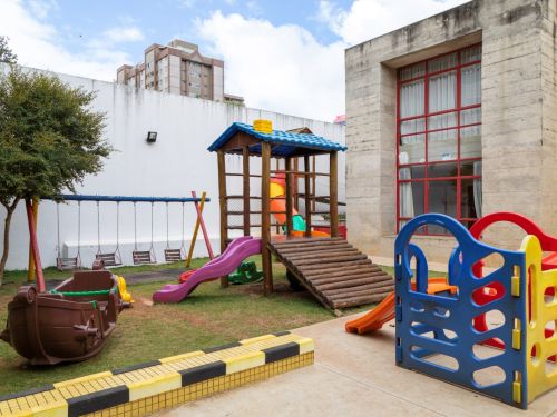 Parquinho em um gramado. Ao centro, um brinquedo de madeira com telhado azul, escorregador roxo e uma pequena ponte de toras. À esquerda, um balanço com vários assentos e um brinquedo em formato de barco. Ao fundo, um muro branco e um prédio de concreto aparente com janelas vermelhas.