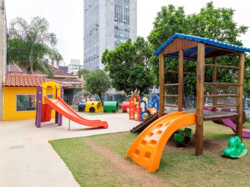 Área de lazer externa com brinquedos sobre grama e cimento. Em primeiro plano, um escorregador laranja e uma casinha de madeira. Ao fundo, um escorregador maior, um túnel de anéis coloridos e uma casinha amarela com telhado de cerâmica. Um prédio alto se destaca ao fundo.