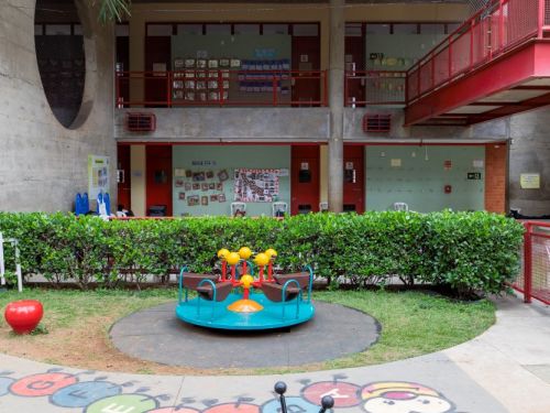 Pátio interno visto de um gramado. No centro, um gira-gira azul e amarelo. Ao fundo, um prédio de dois andares com corredores de grades vermelhas e paredes verde-claras. À esquerda, uma parede de concreto aparente com uma grande abertura circular. Em primeiro plano, um brinquedo de mola.