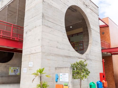 Fachada de um bloco de concreto aparente com uma grande abertura circular no segundo andar. Em frente, um painel informativo e lixeiras coloridas para coleta seletiva. A estrutura tem um design moderno, com corredores de guarda-corpo vermelho visíveis ao fundo.