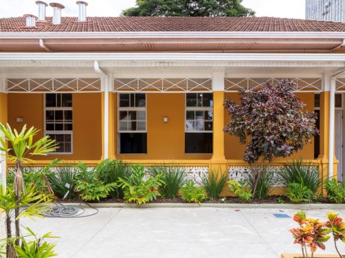 Fachada de uma casa térrea de cor ocre, com telhado de cerâmica e uma varanda coberta com pilares e treliças de madeira branca. Em frente à varanda, um canteiro com plantas de folhagens verdes e roxas. O piso em frente é de cimento claro.
