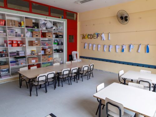 Sala de aula com paredes em tom de creme e piso cinza claro. À esquerda, uma estante branca alta, cheia de caixas e materiais escolares. No centro, uma mesa comprida e retangular com cadeiras infantis. Na parede, um varal com letras do alfabeto e desenhos pendurados em pregadores.