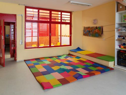 Canto de uma sala de atividades com paredes de cor creme. No chão, um grande tapete emborrachado formado por quadrados de múltiplas cores. Ao fundo, uma ampla janela com esquadrias e persianas vermelhas. À esquerda, uma porta aberta revela outra sala. Uma estante branca está à direita.