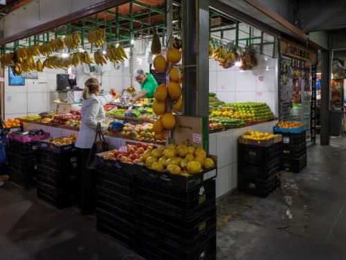 Uma feira com uma banca de frutas em destaque. A banca é coberta por um telhado e tem paredes de azulejos brancos. Em primeiro plano, caixas escuras estão repletas de uma variedade de frutas, como maçãs, pêssegos, laranjas e mangas. Penduradas sobre a banca, cachos de bananas e abacaxis enfeitam o ambiente. Atrás do balcão, um vendedor de touca branca e camiseta verde atende uma cliente.