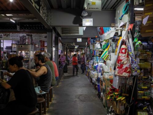 O interior de um mercado em um corredor estreito, com pessoas e bancas dos dois lados. À esquerda, há um balcão com bancos altos onde algumas pessoas estão sentadas, consumindo algo. O ambiente é iluminado por luzes penduradas. À direita, uma banca está cheia de produtos coloridos, como utensílios de cozinha, bolsas, tecidos e artesanato, pendurados em expositores. O corredor é de cimento escuro e, no fundo, outras pessoas e lojas são visíveis.