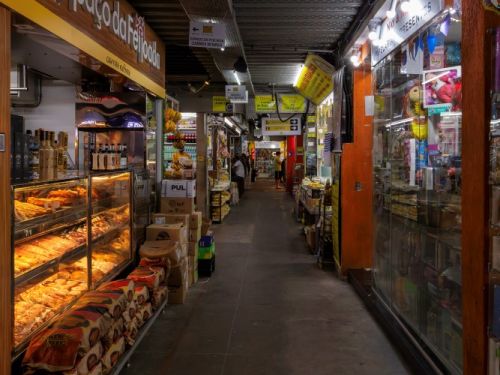 O interior de um corredor estreito e longo em um mercado fechado. O corredor está ladeado por lojas e bancas de produtos, criando uma perspectiva de profundidade. À esquerda, uma loja com a placa "Espaço da Feijoada" tem produtos iluminados, como salgados e carnes, dispostos em balcões de vidro. À direita, há uma banca de doces e produtos embalados, com paredes laranjas e itens expostos em vitrines. O teto está cheio de cabos e estruturas metálicas.